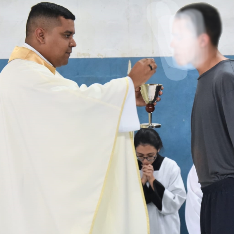Foto mostra à esquerda um padre, um homem negro com batina bege em pé, colocando a hóstia na direção da boca de um adolescente à direita, branco, vestindo camiseta cinza e shorts azul