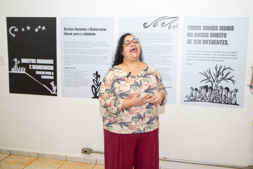 Uma mulher de óculos e blusa floral gesticula e sorri enquanto fala à frente de painéis sobre cidadania e direitos humanos.