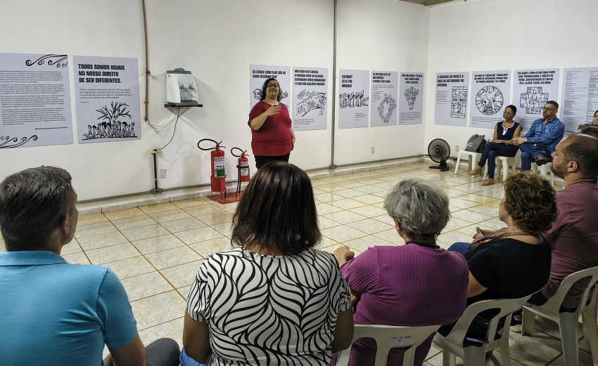 Uma mulher de blusa vermelha fala para uma plateia sentada em semicírculo em uma sala com cartazes educativos nas paredes.