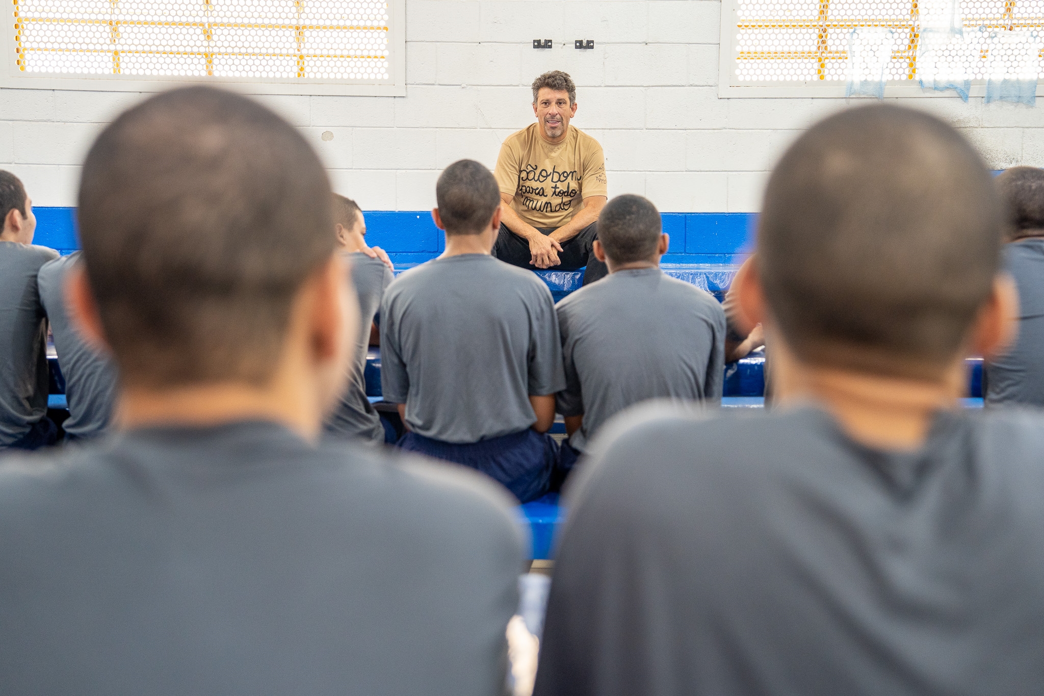 Milhem Cortaz está sentado de frente em um banco azul, conversando com um grupo de jovens sentados de costas que o observam atentamente.