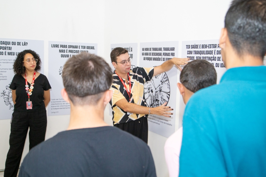 Um educador de camisa estampada explica o conteúdo dos painéis na parede para um grupo de jovens vistos de costas.
