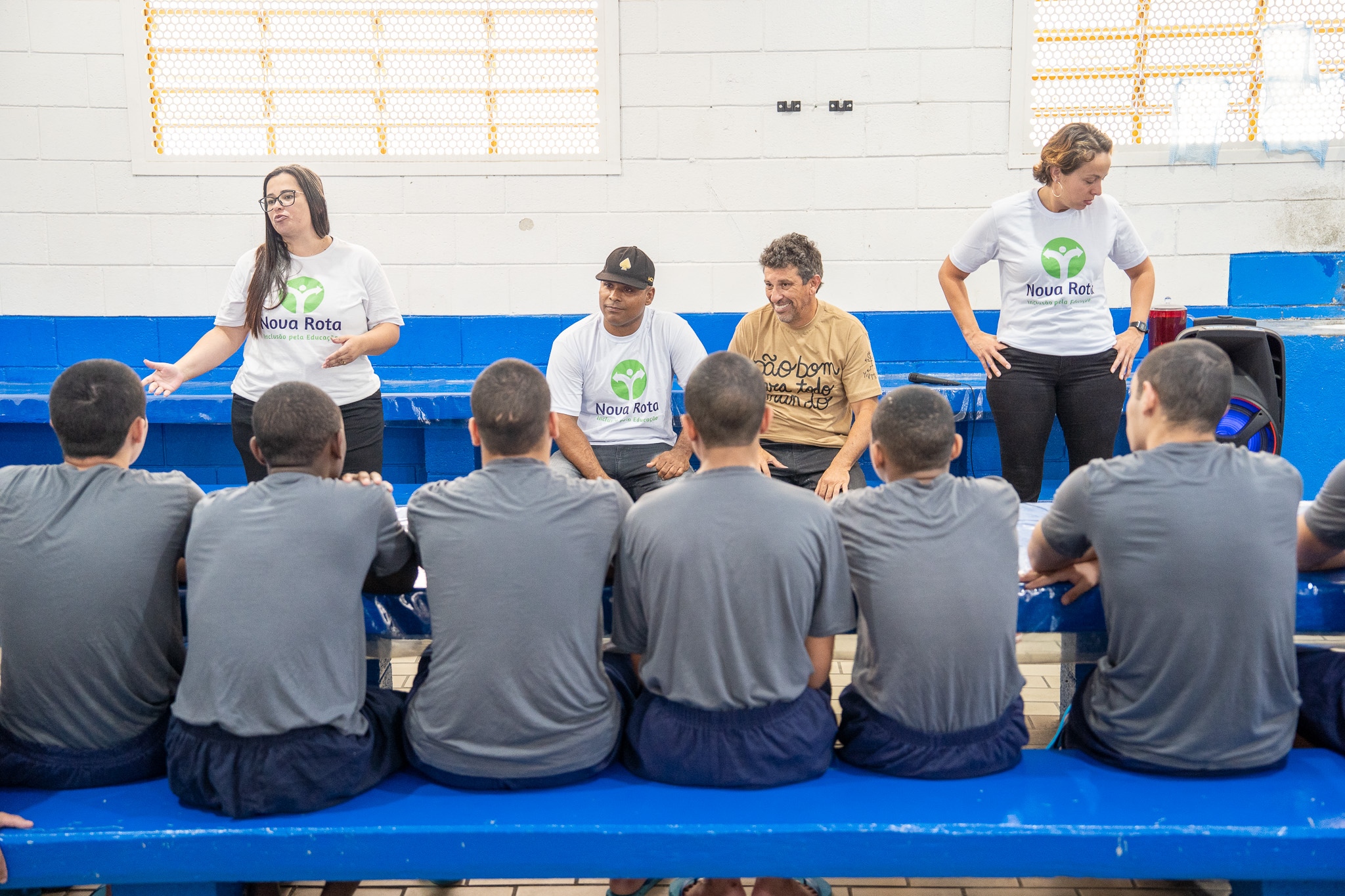 Duas mulheres em pé e dois homens sentados conversam com um grupo de jovens sentados de costas; uma delas gesticula enquanto o grupo usa camisetas com o logo "Nova Rota".