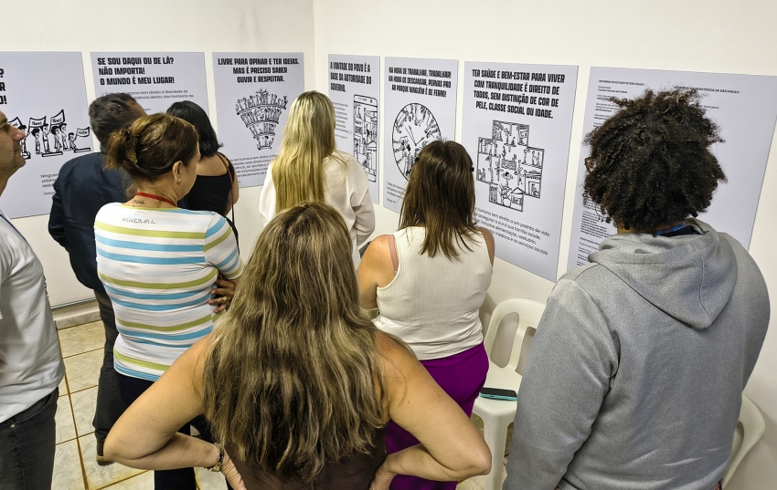 Vista superior de um grupo de pessoas de costas, observando atentamente diversos painéis educativos fixados em uma parede branca.