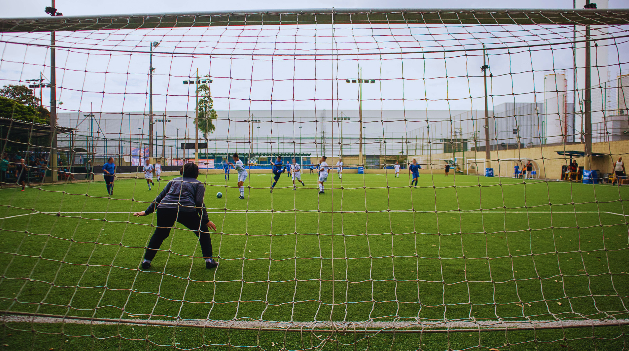  Vista do gol mostra jogadoras em ação durante partida de futebol society, com goleira posicionada para defesa.