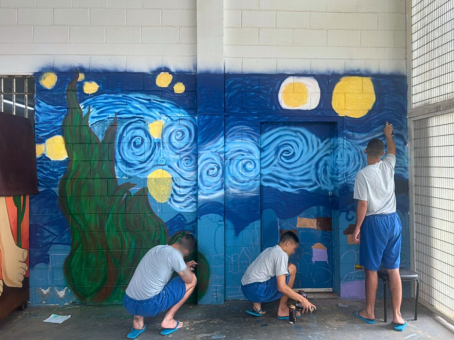 dolescentes pintam mural em parede interna de centro socioeducativo. A arte, em andamento, é inspirada na obra “Noite Estrelada”, de Vincent van Gogh, com tons de azul e amarelo. Um jovem pinta em pé e dois trabalham agachados com spray.