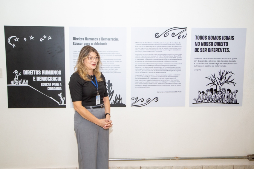 Uma mulher loira de óculos e camiseta preta sorri e fala em frente a quatro painéis brancos e pretos sobre democracia.