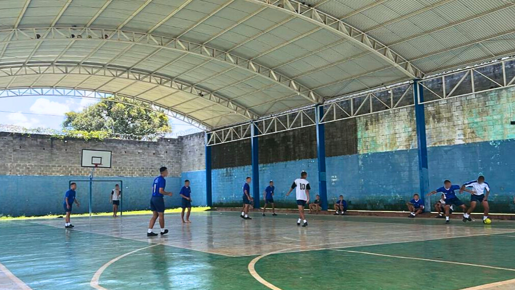 Grupo de adolescentes joga futsal em quadra coberta, enquanto outros observam sentados ao lado da parede azul e branca.
