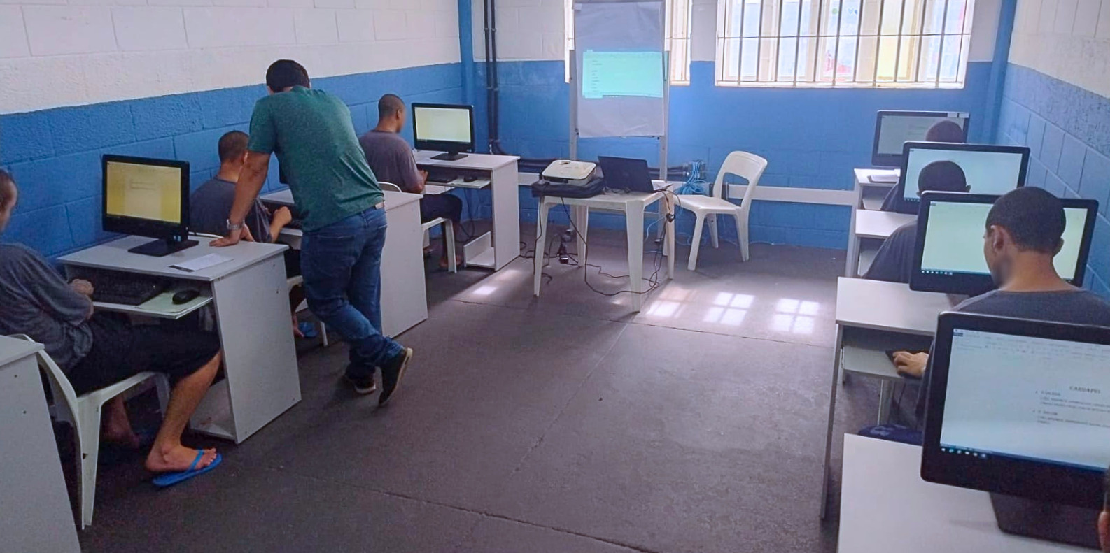 Sala de informática com adolescentes sentados em computadores, realizando atividades enquanto um educador orienta um dos participantes.