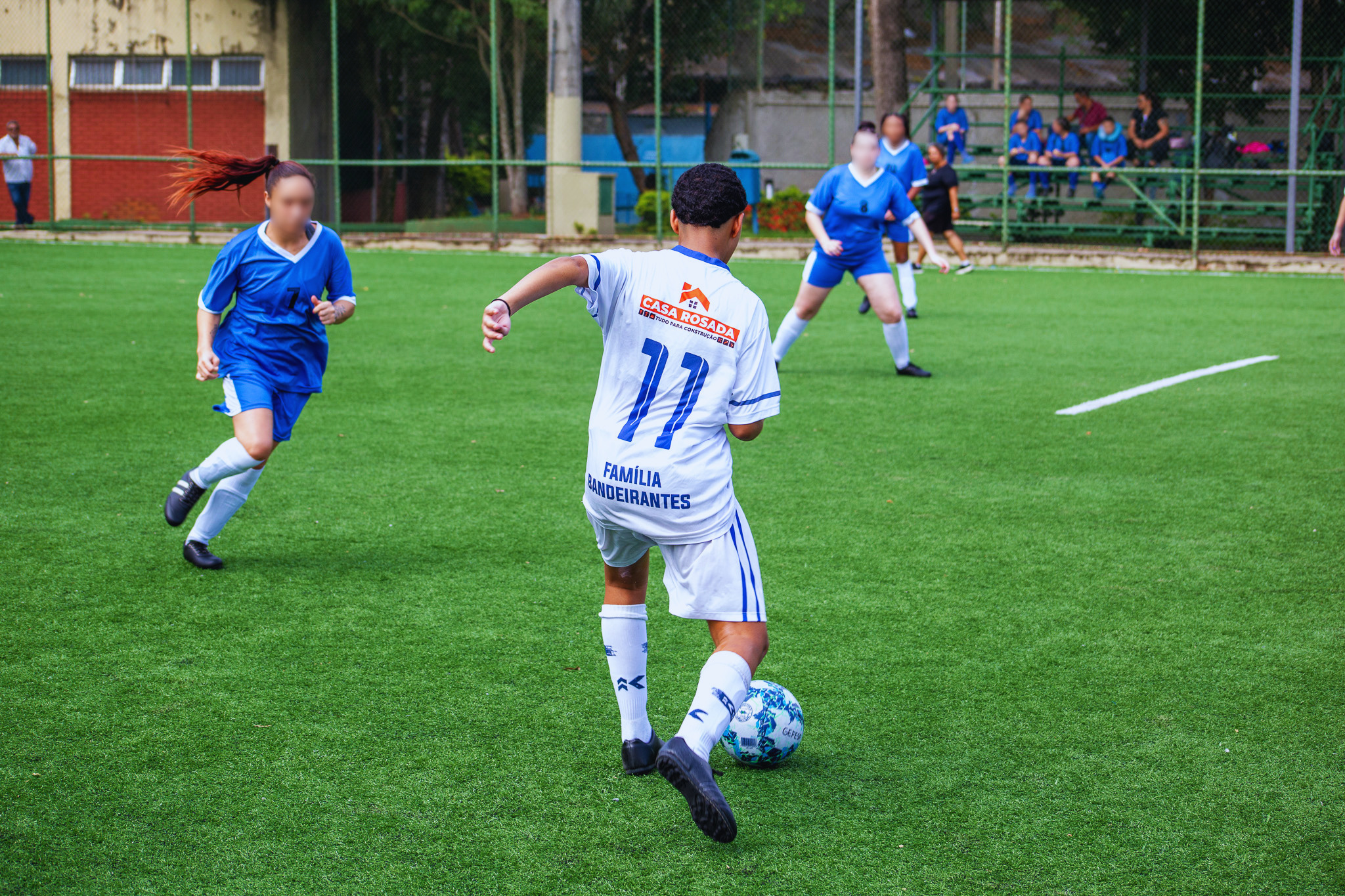 Jogadora conduz a bola em campo enquanto é marcada por adversária, com outras atletas ao fundo acompanhando a jogada.