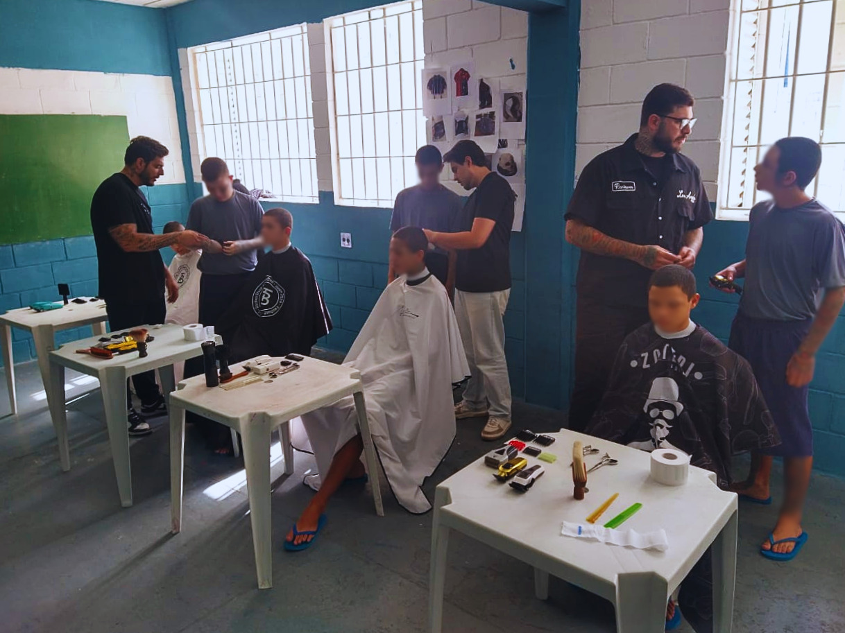 Vários adolescentes participam de cortes de cabelo simultâneos em sala interna. Mesas com equipamentos organizados.